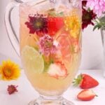 A glass pitcher filled with a refreshing drink containing ice, sliced strawberries, lime, and colorful edible flowers, surrounded by flowers, a strawberry, and a lime wedge on a white surface.