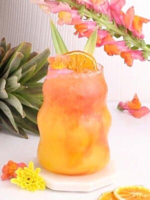 A colorful tropical mocktail drink with orange and pink hues is served in a curvy glass, garnished with a dried orange slice and pineapple leaves. Flowers and a pineapple are in the background, adding a vibrant, summery feel.