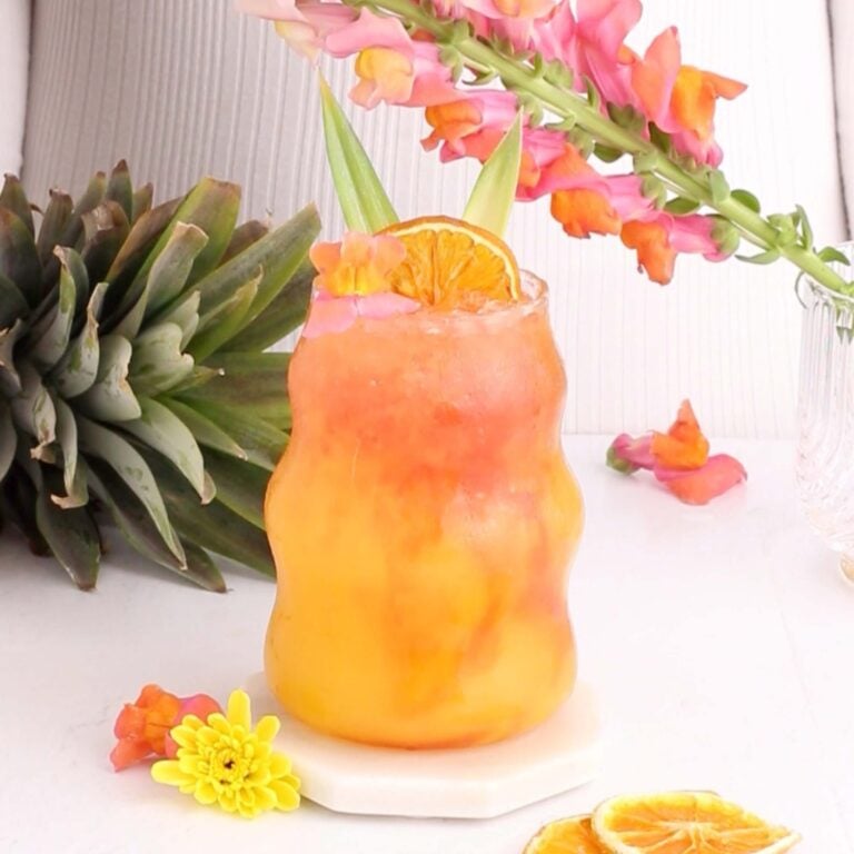 A colorful tropical mocktail drink with orange and pink hues is served in a curvy glass, garnished with a dried orange slice and pineapple leaves. Flowers and a pineapple are in the background, adding a vibrant, summery feel.