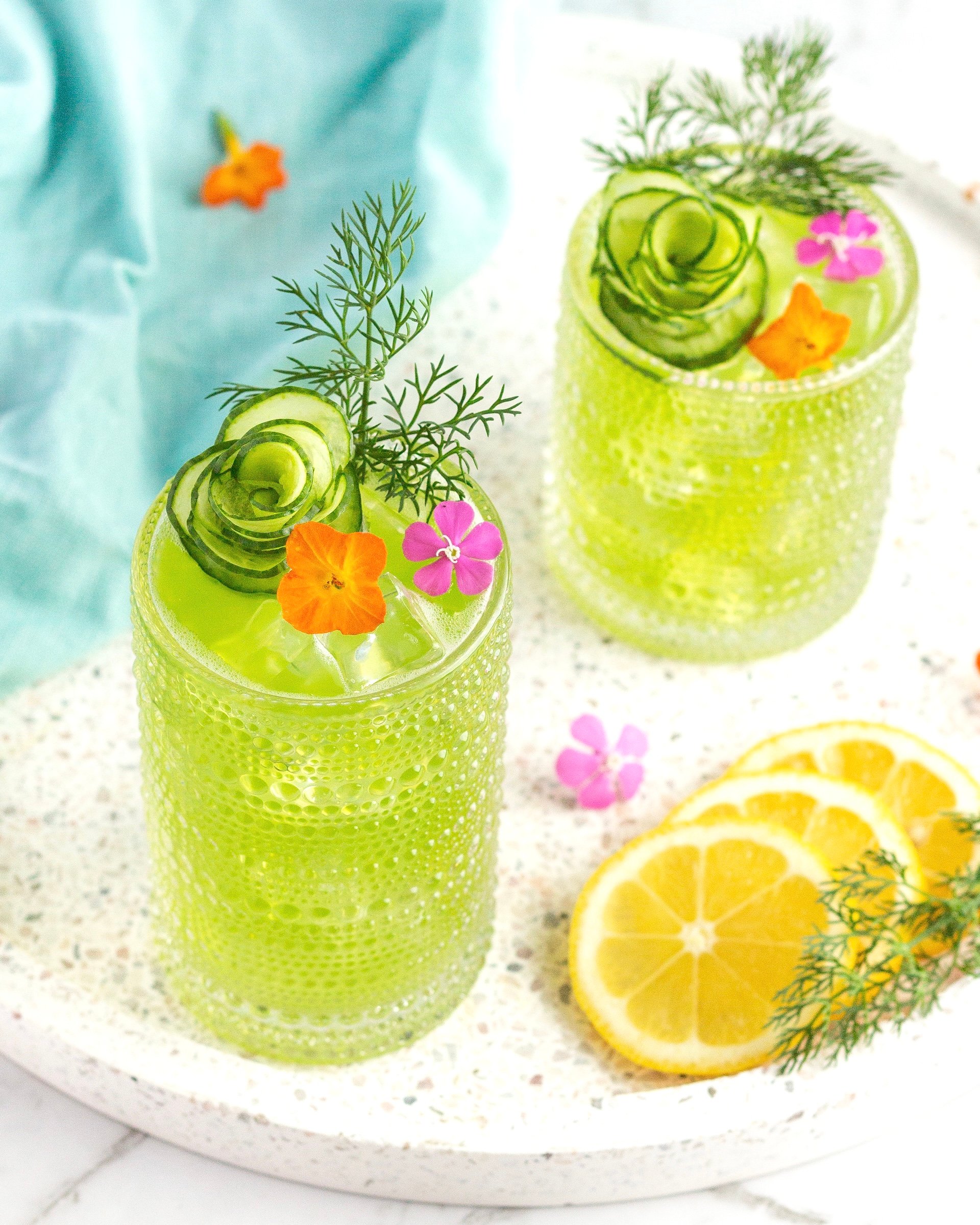 Two green drinks in textured glasses are garnished with cucumber spirals, dill, and colorful edible flowers. Lemon slices and extra flowers decorate the white speckled tray they sit on. A light blue cloth is in the background.