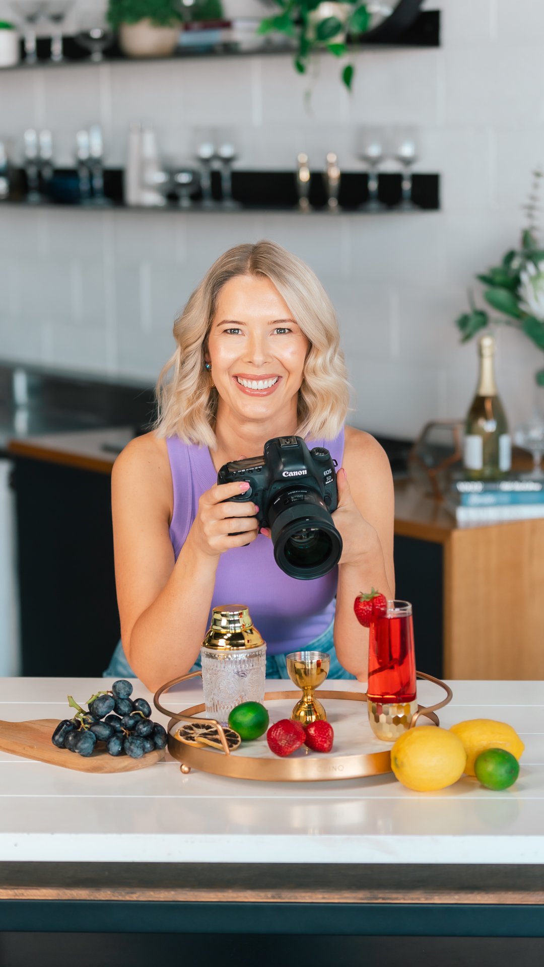 A smiling woman with blonde hair holds a camera in a modern kitchen, standing behind a counter with grapes, lemons, limes, strawberries, a drink, and bar tools. Shelves with glassware and a wine bottle are in the background.