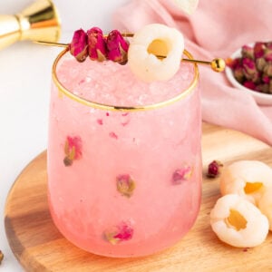 A pink lychee mocktail with ice and rose petals in a glass with a gold rim, garnished with lychee and dried rosebuds on a pick, placed on a wooden board with fresh lychees nearby.