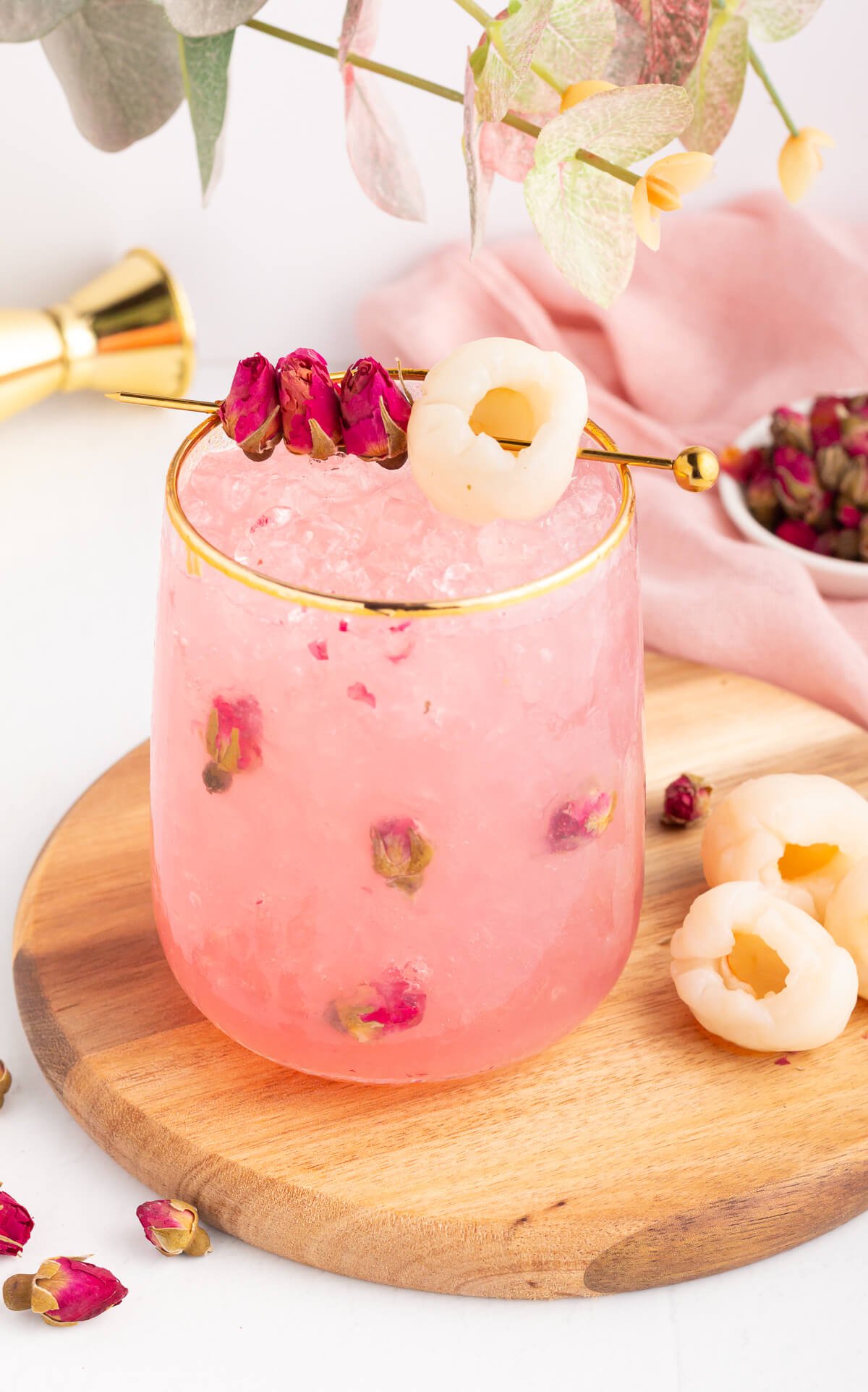 A pink lychee mocktail with ice and rose petals in a glass with a gold rim, garnished with lychee and dried rosebuds on a pick, placed on a wooden board with fresh lychees nearby.