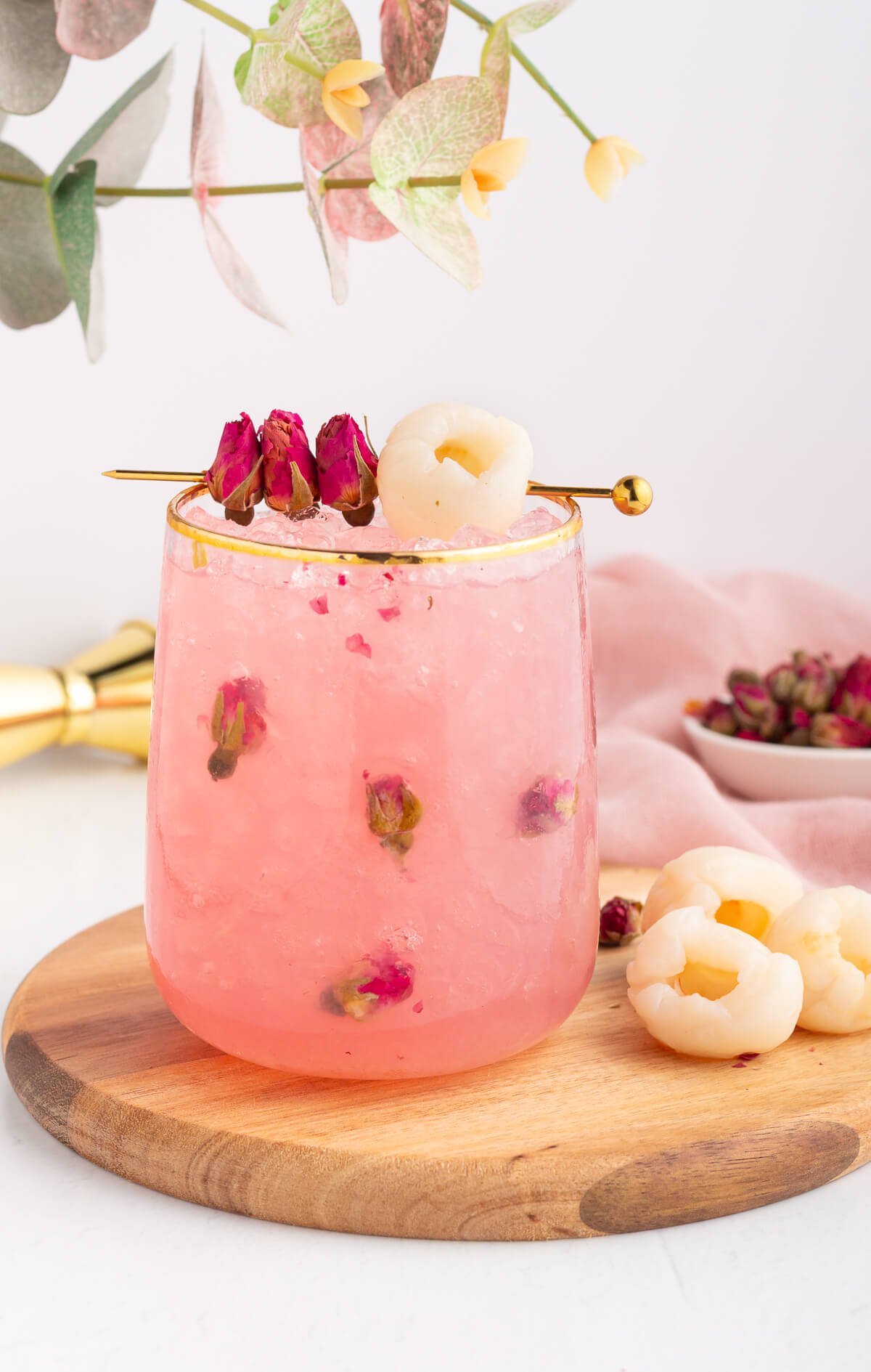 A pink lychee drink in a glass with crushed ice, rose petals, and a gold rim, garnished with dried rosebuds and a lychee on a pick, sits on a wooden board with fresh lychees and rosebuds beside it.