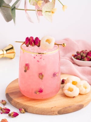 A pink lychee drink with rosebuds in a glass on a wooden board, garnished with lychee fruit and dried rosebuds on a cocktail pick, with a small bowl of rosebuds and a pink cloth in the background.