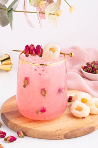 A pink lychee drink with rosebuds in a glass on a wooden board, garnished with lychee fruit and dried rosebuds on a cocktail pick, with a small bowl of rosebuds and a pink cloth in the background.