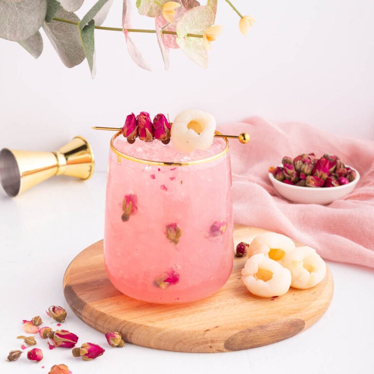 A pink lychee drink with rosebuds in a glass on a wooden board, garnished with lychee fruit and dried rosebuds on a cocktail pick, with a small bowl of rosebuds and a pink cloth in the background.