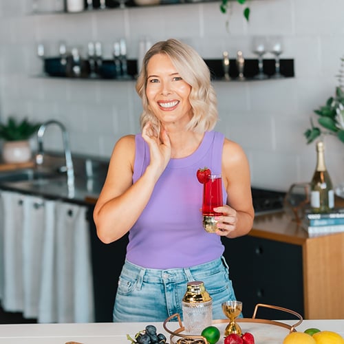 A smiling woman with blonde hair in a lavender sleeveless top holds a red drink with a raspberry garnish in a modern kitchen. Various fruits and cocktail-making items are on the counter in front of her.