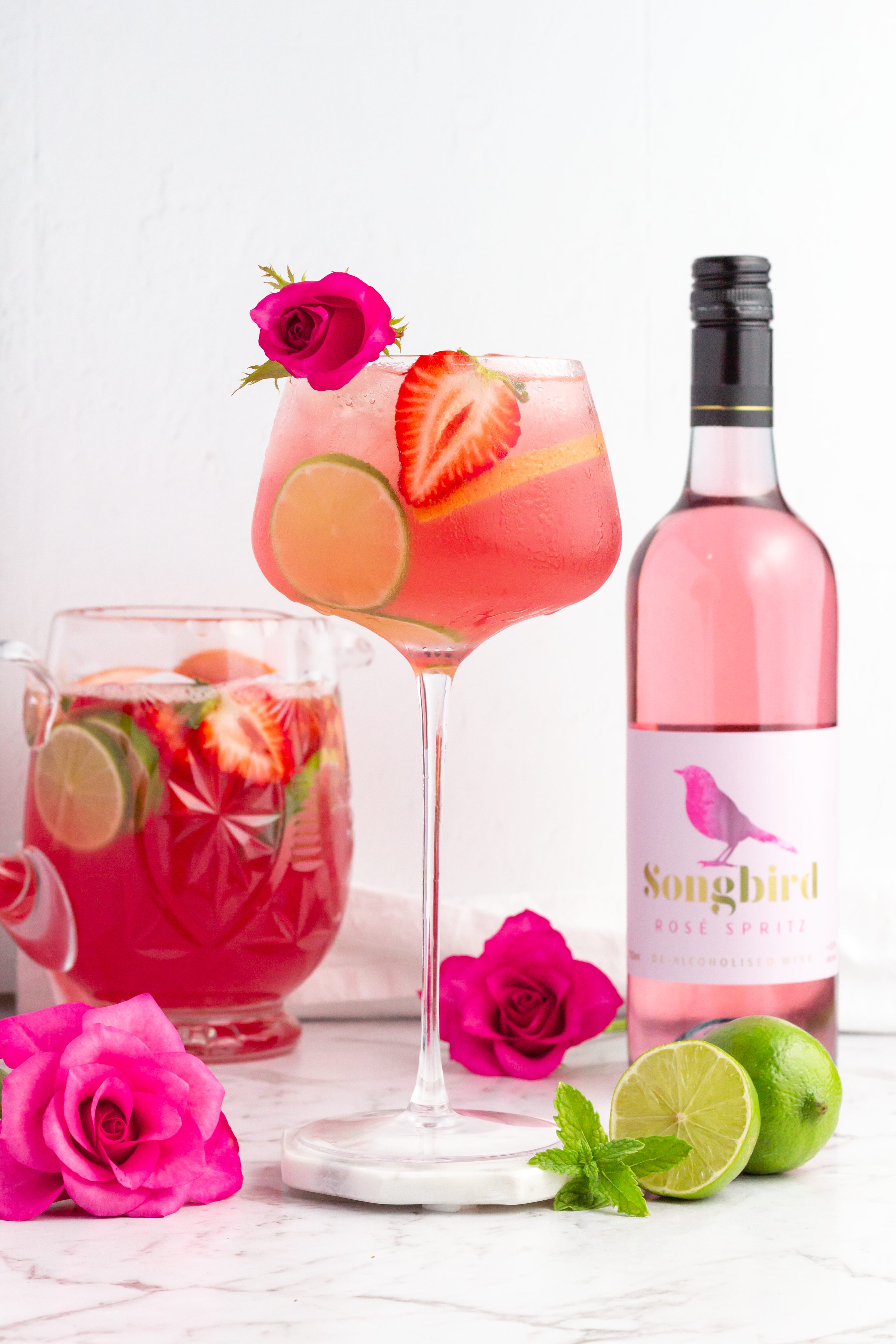 A glass of sparkling red drink garnished with a blood orange slice and red flowers sits on a coaster next to a bottle of Edenvale non-alcoholic wine and more red flowers on a white background.