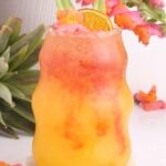 A vibrant tropical mocktail drink with orange and red layers in a curvy glass, garnished with a dried orange slice and green leaves. Bright flowers and a pineapple are in the background, creating a summery, festive vibe.