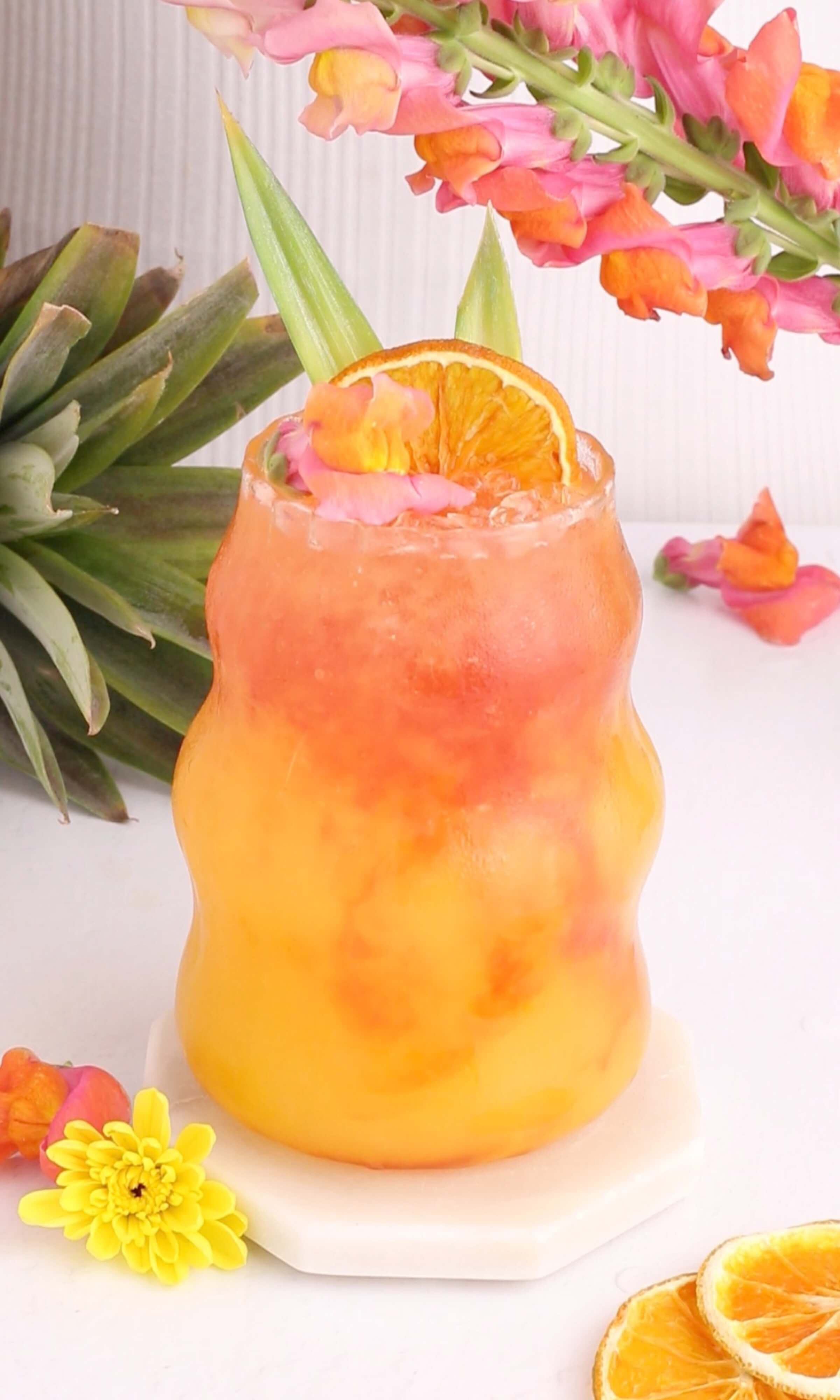 A vibrant, layered orange and pink tropical drink garnished with a dried orange slice, pineapple leaves, and pink flowers, sits on a white coaster surrounded by colorful flowers and a pineapple.