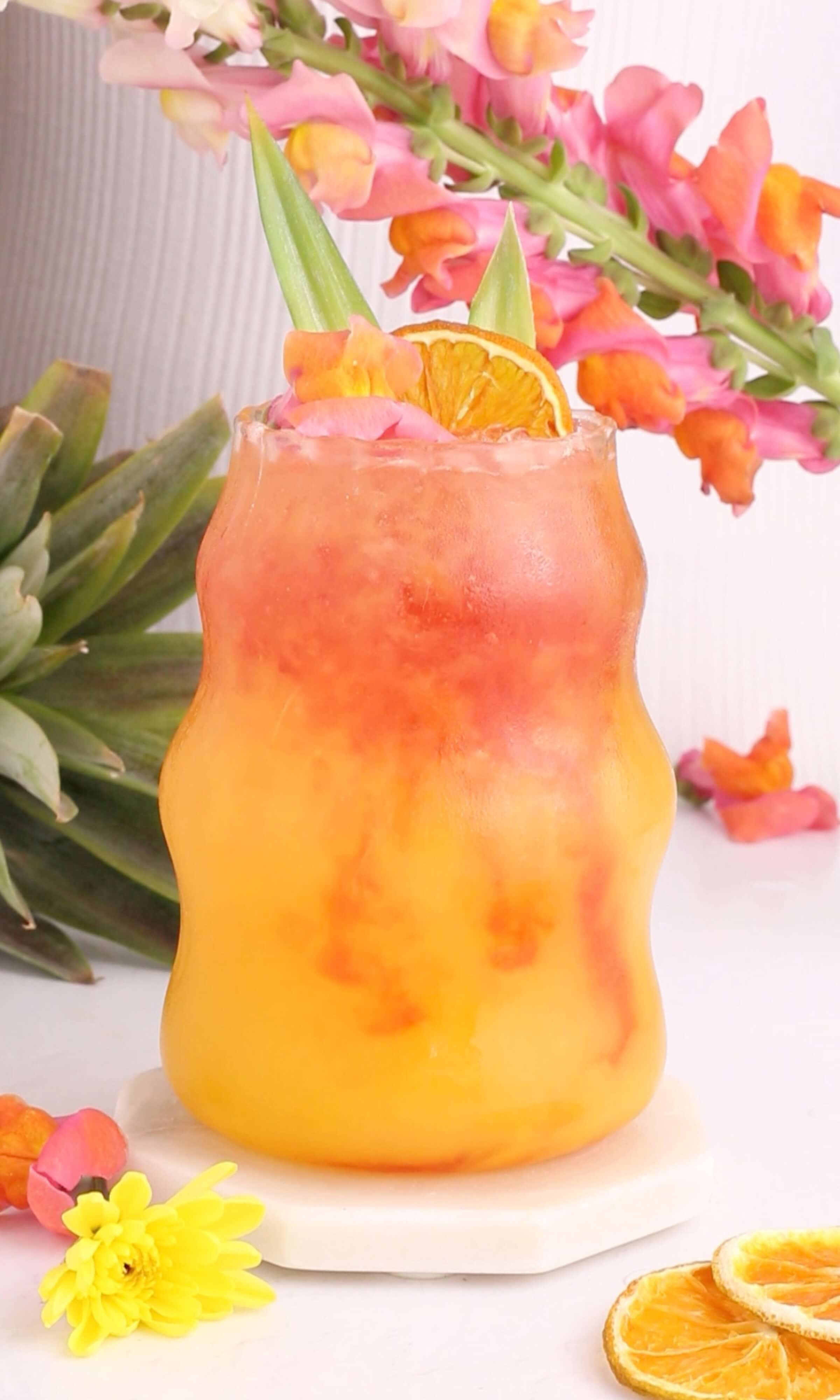 A vibrant tropical mocktail drink with orange and red layers in a curvy glass, garnished with a dried orange slice and green leaves. Bright flowers and a pineapple are in the background, creating a summery, festive vibe.