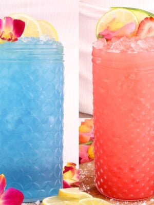 Two tall textured glasses, one with a bright blue drink garnished with lemon slices and a flower, the other with a pink drink topped with crushed ice, lime slices, strawberries, and a flower. Both are surrounded by fruit and petals.