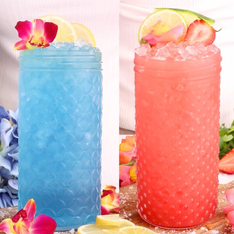 Two tall textured glasses, one with a bright blue drink garnished with lemon slices and a flower, the other with a pink drink topped with crushed ice, lime slices, strawberries, and a flower. Both are surrounded by fruit and petals.