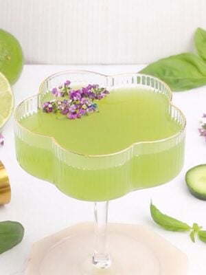 A green cocktail in a flower-shaped glass garnished with small purple flowers. Surrounding the glass are a sliced lime, cucumber slice, basil leaves, a gold jigger, and more flowers on a white surface.