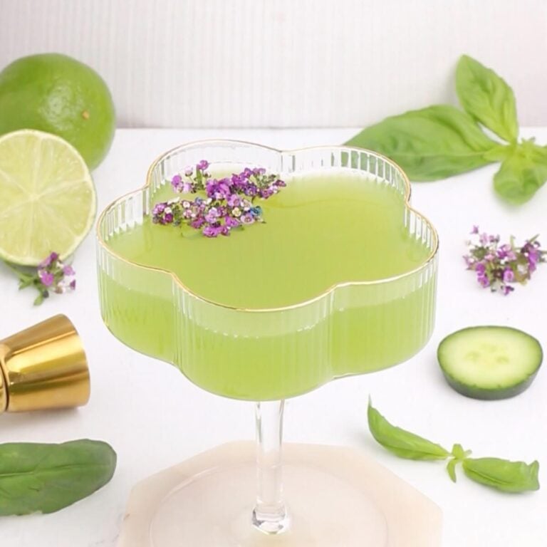 A green cocktail in a flower-shaped glass garnished with small purple flowers. Surrounding the glass are a sliced lime, cucumber slice, basil leaves, a gold jigger, and more flowers on a white surface.