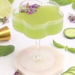 A pale green cocktail in a flower-shaped glass, garnished with small purple flowers. Fresh basil leaves, cucumber slices, a gold spoon, and a halved lime are arranged around the glass on a white surface.