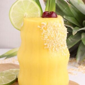 A yellow tropical smoothie garnished with shredded coconut, a lime wheel, a cherry, and pineapple leaves, served in a curvy glass on a wooden board with lime slices and a pineapple in the background.