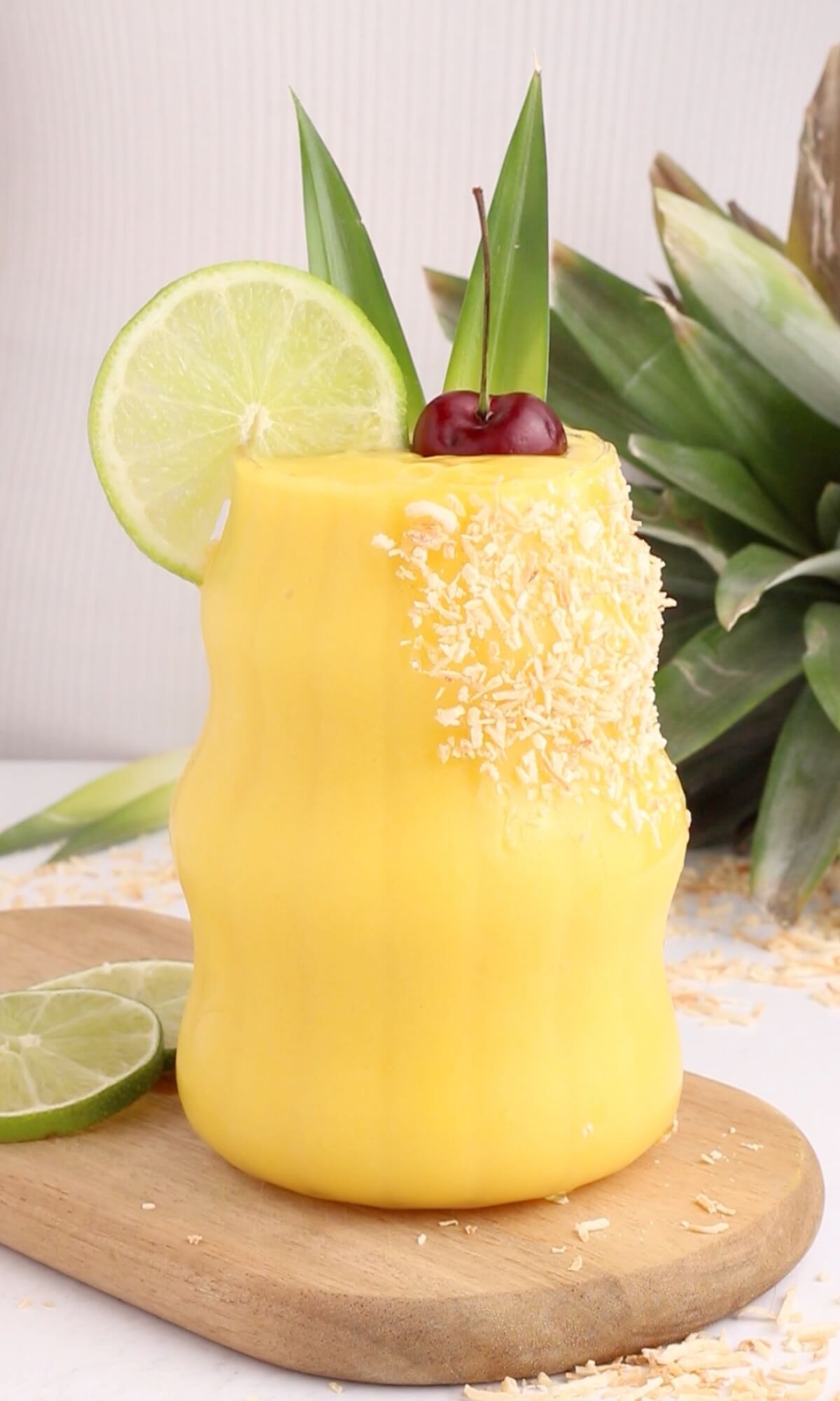 A yellow tropical smoothie garnished with shredded coconut, a lime wheel, a cherry, and pineapple leaves, served in a curvy glass on a wooden board with lime slices and a pineapple in the background.