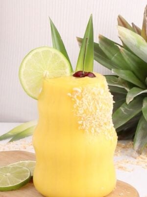 A yellow non-alcoholic mango colada garnished with shredded coconut, a lime slice, pineapple leaves, and a cherry sits on a wooden board beside lime slices and a fresh pineapple.