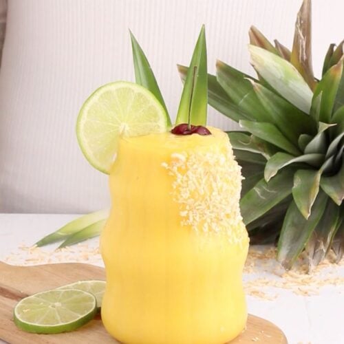 A yellow non-alcoholic mango colada garnished with shredded coconut, a lime slice, pineapple leaves, and a cherry sits on a wooden board beside lime slices and a fresh pineapple.