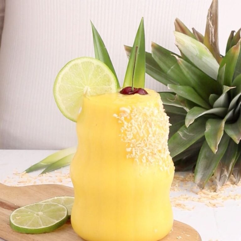 A yellow non-alcoholic mango colada garnished with shredded coconut, a lime slice, pineapple leaves, and a cherry sits on a wooden board beside lime slices and a fresh pineapple.