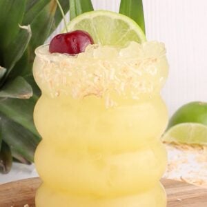 A tropical pineapple punch in a wavy glass, topped with a lime wheel, cherry, and pineapple leaves. Toasted coconut flakes rim the glass. Pineapple and lime slices are in the background on a wooden surface.