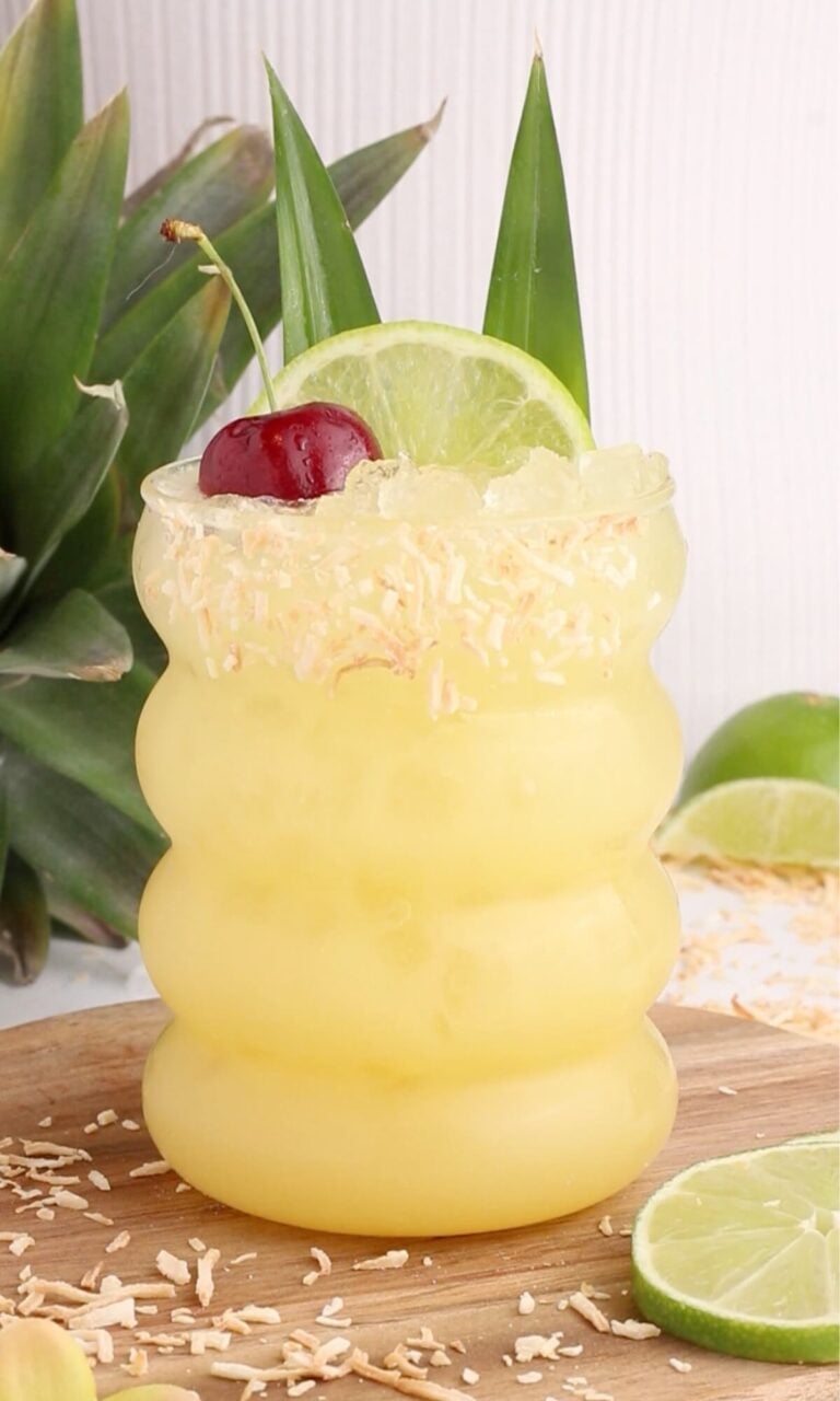 A tropical pineapple punch in a wavy glass, topped with a lime wheel, cherry, and pineapple leaves. Toasted coconut flakes rim the glass. Pineapple and lime slices are in the background on a wooden surface.