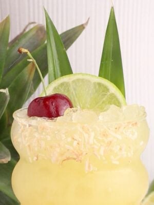 A yellow tropical drink with crushed ice in a curved glass, garnished with toasted coconut on the rim, a lime slice, a cherry, and two pineapple leaves. A fresh pineapple and lime are in the background.