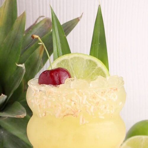 A yellow tropical drink with crushed ice in a curved glass, garnished with toasted coconut on the rim, a lime slice, a cherry, and two pineapple leaves. A fresh pineapple and lime are in the background.
