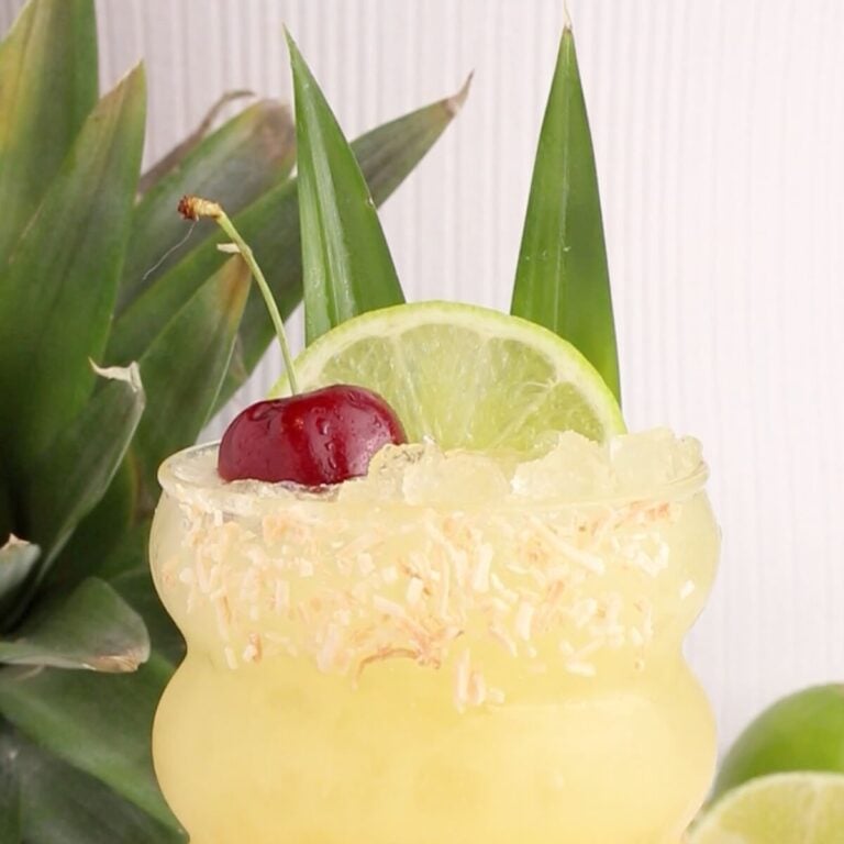 A yellow tropical drink with crushed ice in a curved glass, garnished with toasted coconut on the rim, a lime slice, a cherry, and two pineapple leaves. A fresh pineapple and lime are in the background.