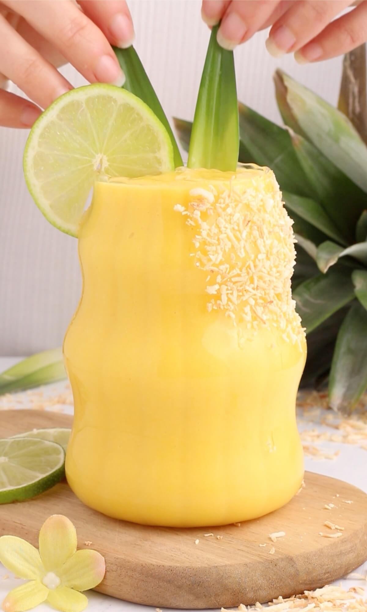A creamy yellow tropical smoothie in a wavy glass, garnished with a lime slice, shredded coconut, and two pandan leaves. Hands adjust the leaves. Pineapple and lime are in the background on a wooden board.