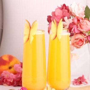 Two tall glasses of orange juice are garnished with butterfly-shaped apple slices. Pink flowers and a peach sit nearby, and a person in a light ribbed shirt stands behind them.