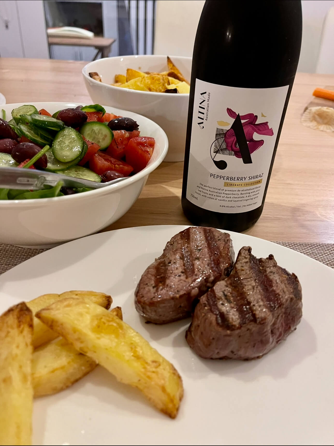 A plate with grilled steak, potato wedges, and a salad. A bowl of salad with tomatoes, cucumber, and olives, and a bottle of Pepperberry Shiraz red wine are on the table.