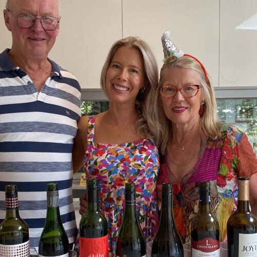 Three people stand smiling behind a counter lined with bottles of non-alcoholic wine. The man wears glasses and a striped shirt; the young woman wears a colorful dress; the older woman wears glasses, a red dress, and a festive party hat.