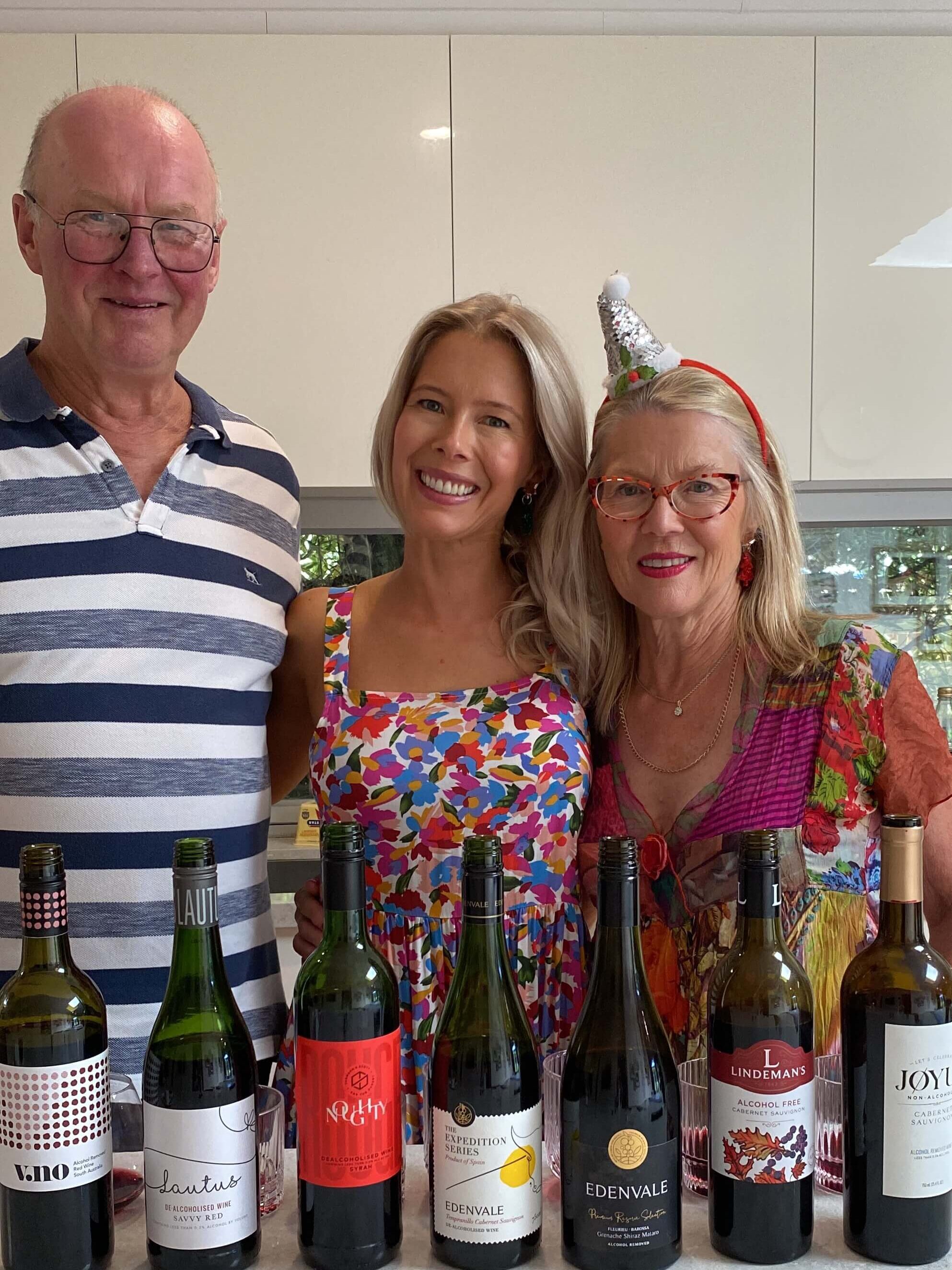 Three people stand smiling behind a counter lined with bottles of non-alcoholic wine. The man wears glasses and a striped shirt; the young woman wears a colorful dress; the older woman wears glasses, a red dress, and a festive party hat.