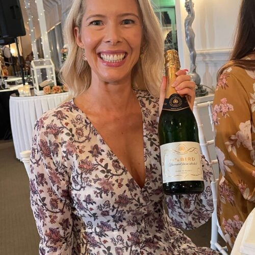 A smiling woman in a floral dress sits at a table, holding a bottle of non-alcoholic champagne at an indoor event. There is a lamp behind her and another person is partially visible on the right.