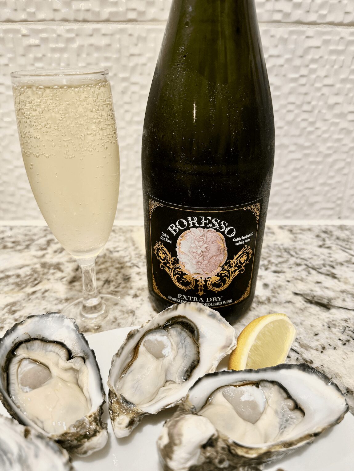 A bottle of Boresso Extra Dry alcohol-free sparkling wine next to a filled champagne flute, served with fresh oysters on a plate and a lemon wedge, set on a marble countertop.