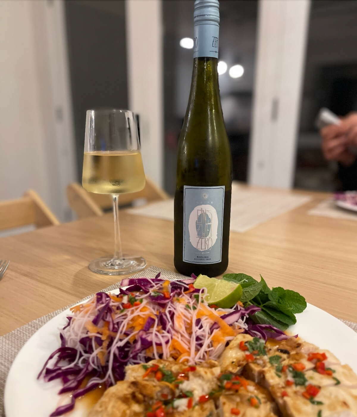 A plate of grilled chicken with chili, served with a colorful salad of cabbage, carrots, and herbs sits on a table next to a glass and bottle of white wine. The background shows a modern dining area.