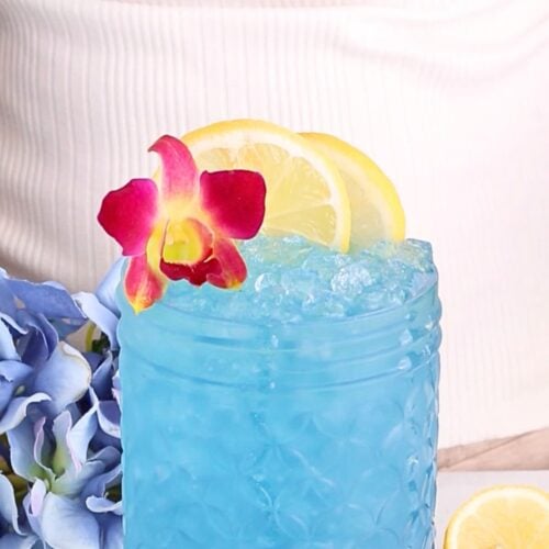 A vibrant blue homemade electrolyte drink in a textured glass, topped with crushed ice, lemon slices, and a pink orchid flower. Blue hydrangea flowers and a lemon wedge sit nearby against a light background.