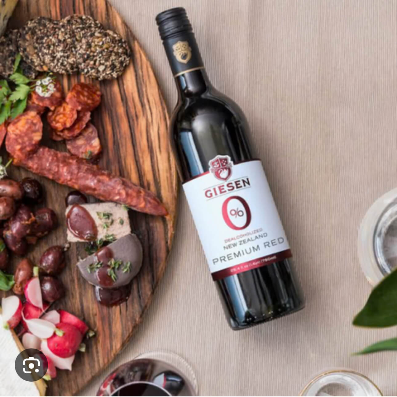 A bottle of Giesen Premium Red wine lies on a table next to a charcuterie board with cured meats, nuts, olives, radishes, crackers, and herbs, alongside two glasses of wine.
