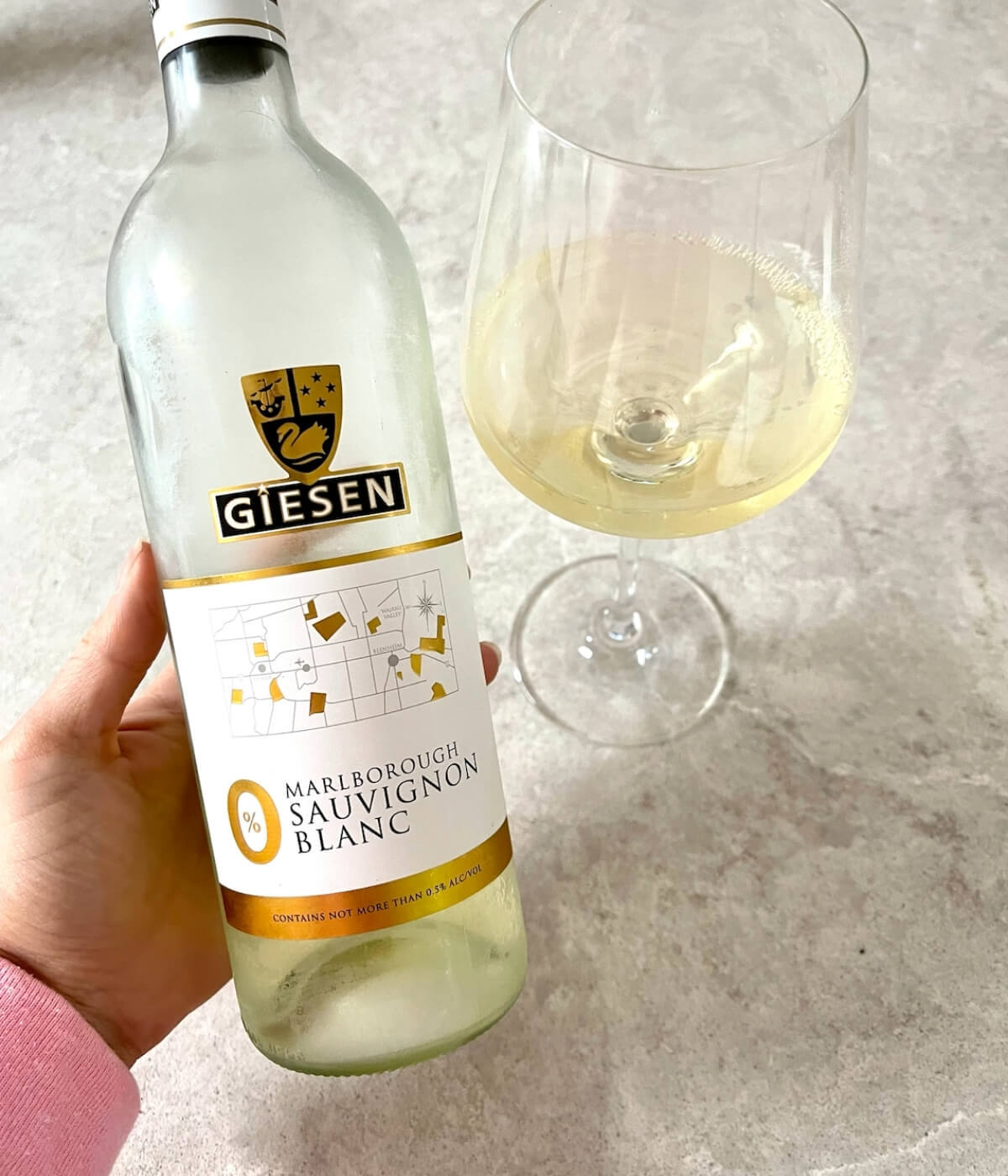 A hand holds a bottle of Giesen Marlborough Sauvignon Blanc 0% beside a glass half-filled with the white wine, set on a light-colored surface.