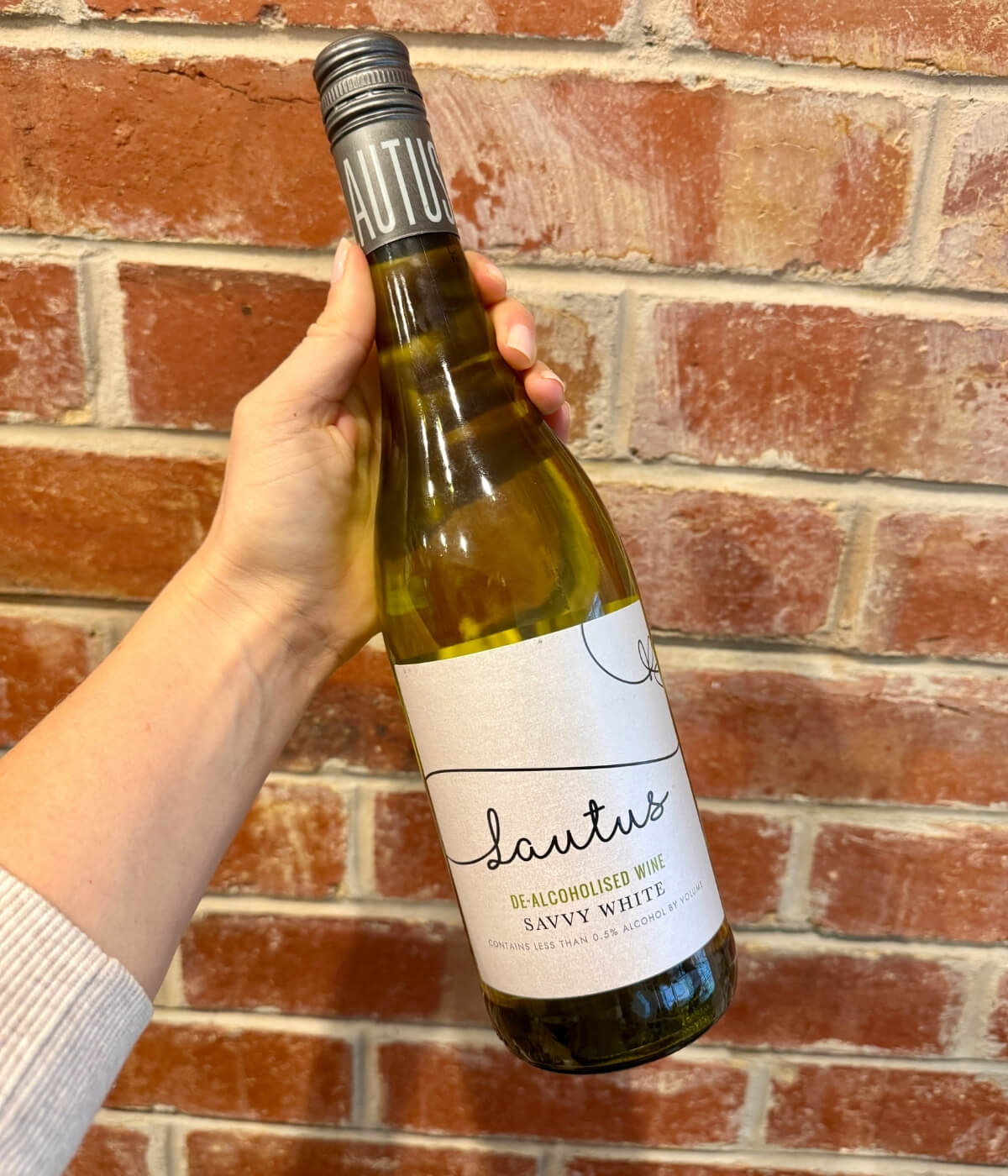 A hand holds a bottle of Lautus De-Alcoholised Wine Savvy White in front of a red brick wall. The bottle has a light-colored label with black cursive and printed text.