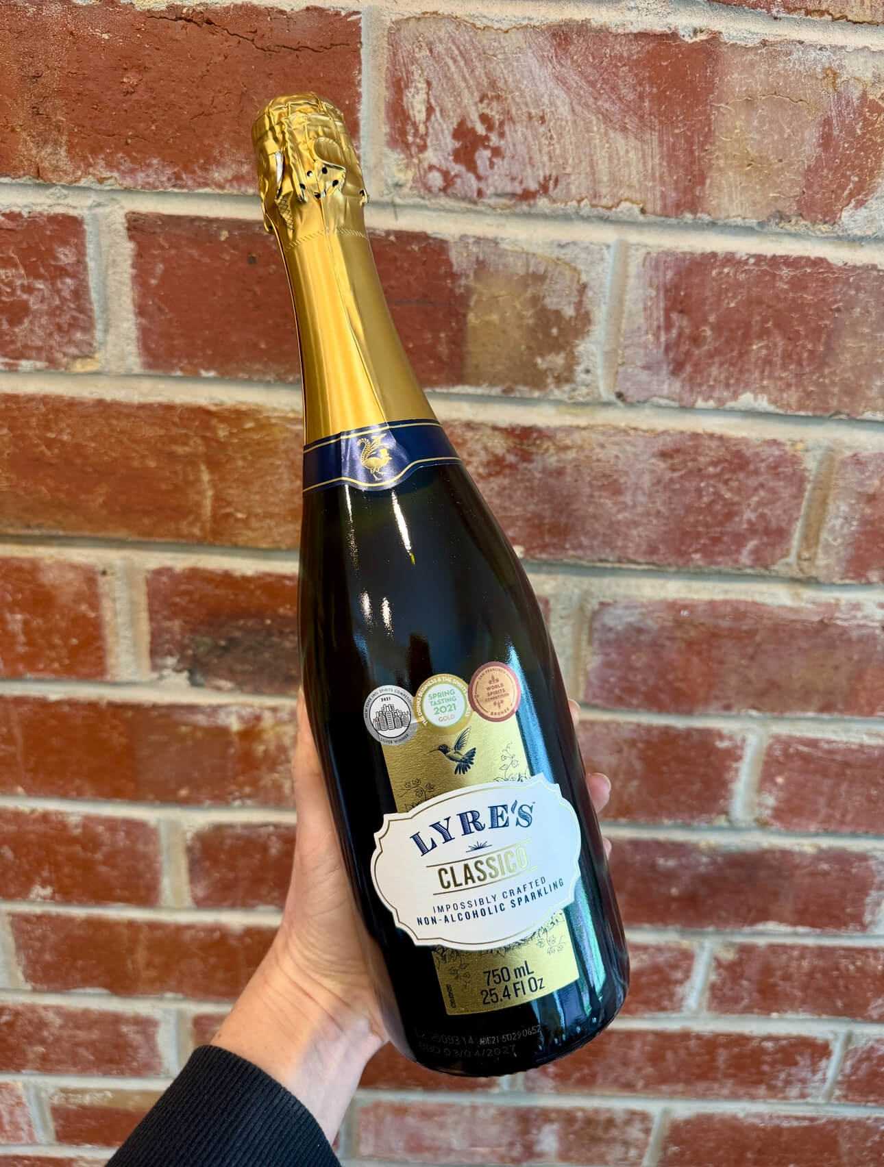 A hand holds a bottle of Lyreโs Classico non-alcoholic sparkling wine in front of a red brick wall. The bottle has gold foil on the top and several award stickers on the label.