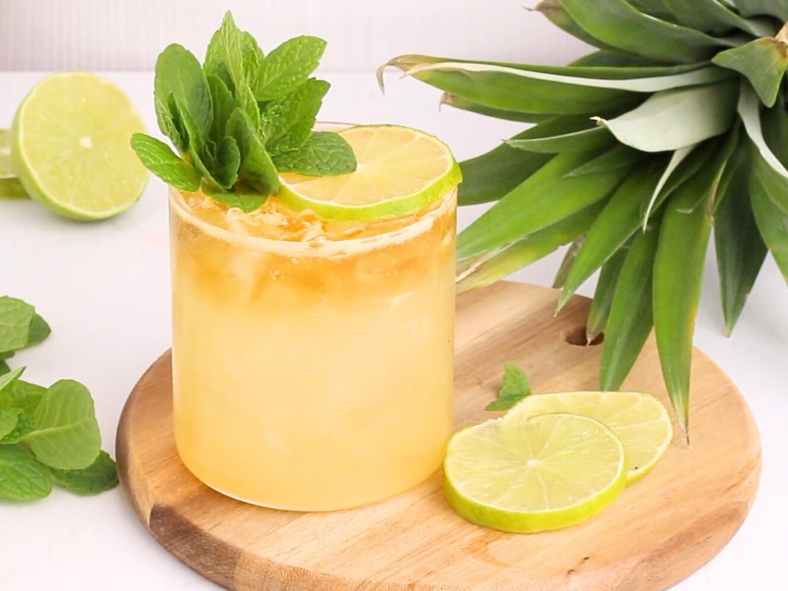 A refreshing yellow drink with ice, garnished with a lime wheel and fresh mint, sits on a wooden board next to lime slices, mint leaves, and part of a pineapple.