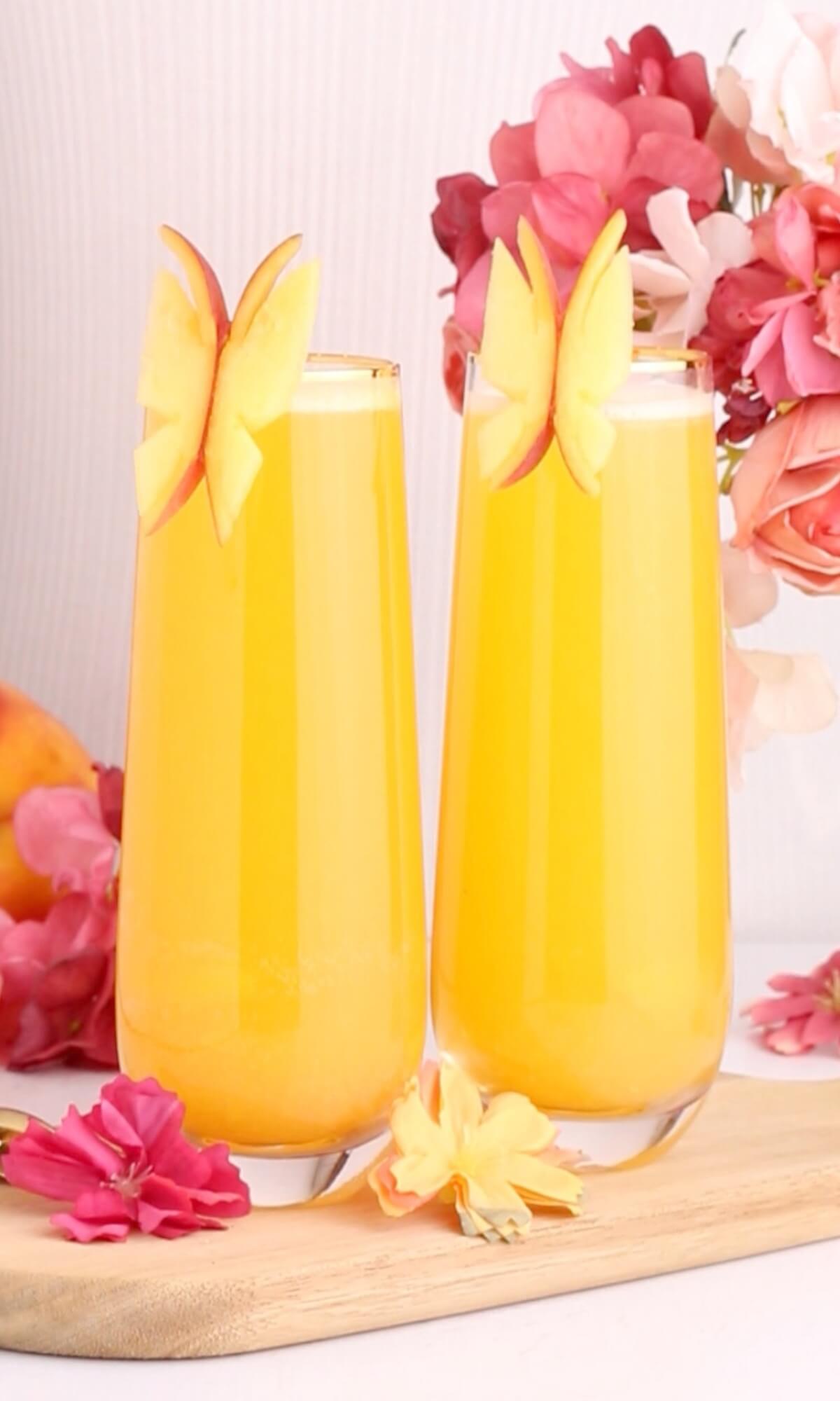 Two tall glasses of orange juice are garnished with butterfly-shaped apple slices on the rims, set on a wooden board with pink and yellow flowers around them and a soft floral background.