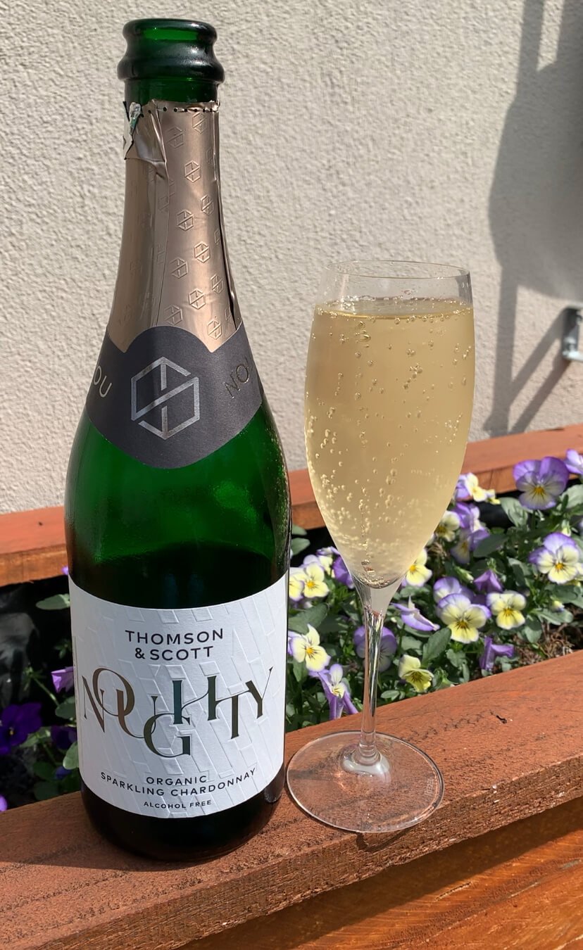 A bottle of Thomson & Scott Noughty organic sparkling chardonnay and a filled flute glass sit on a wooden ledge with blooming purple and yellow flowers in the background.