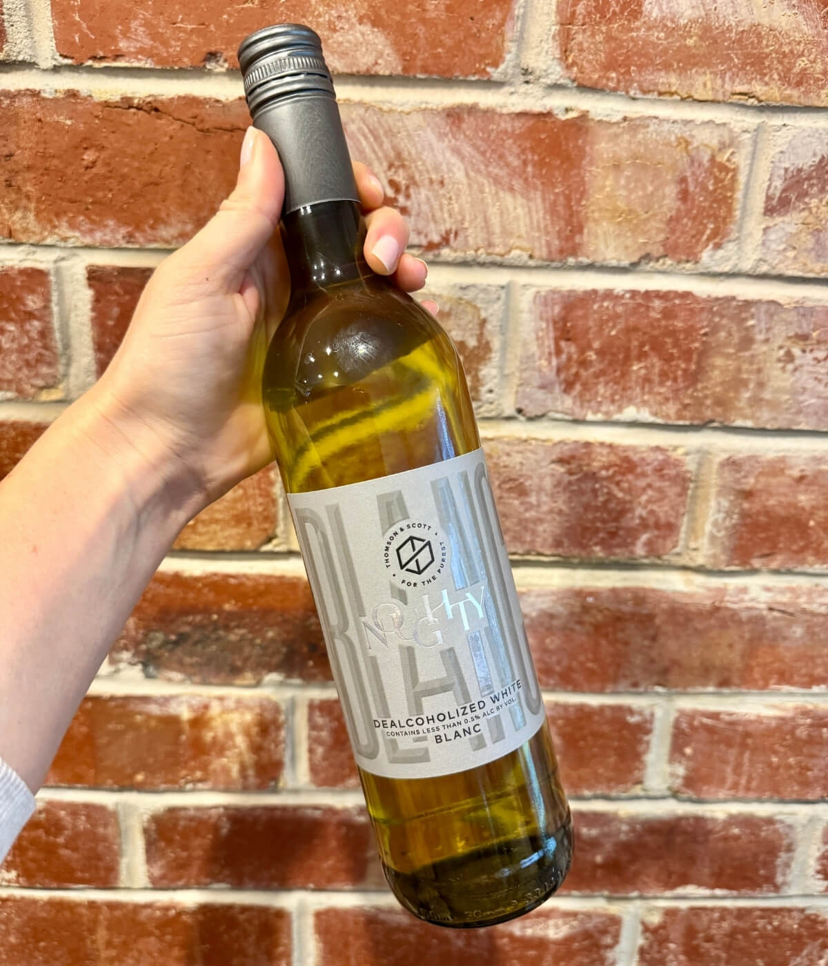 A hand holds a bottle of Noughty alcohol-free white wine in front of a red brick wall. The wine bottle has a silver cap and a light gray label with modern design elements.
