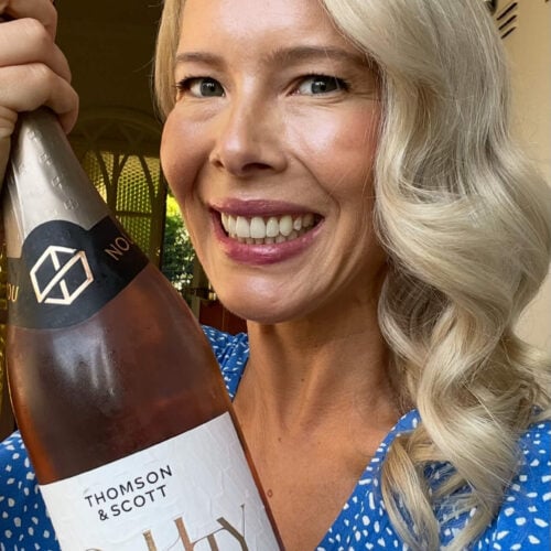 A smiling woman with long blond hair wearing a blue polka dot dress holds up a bottle of Thomson & Scott Noughty organic sparkling rosé. She stands indoors in front of an open door.