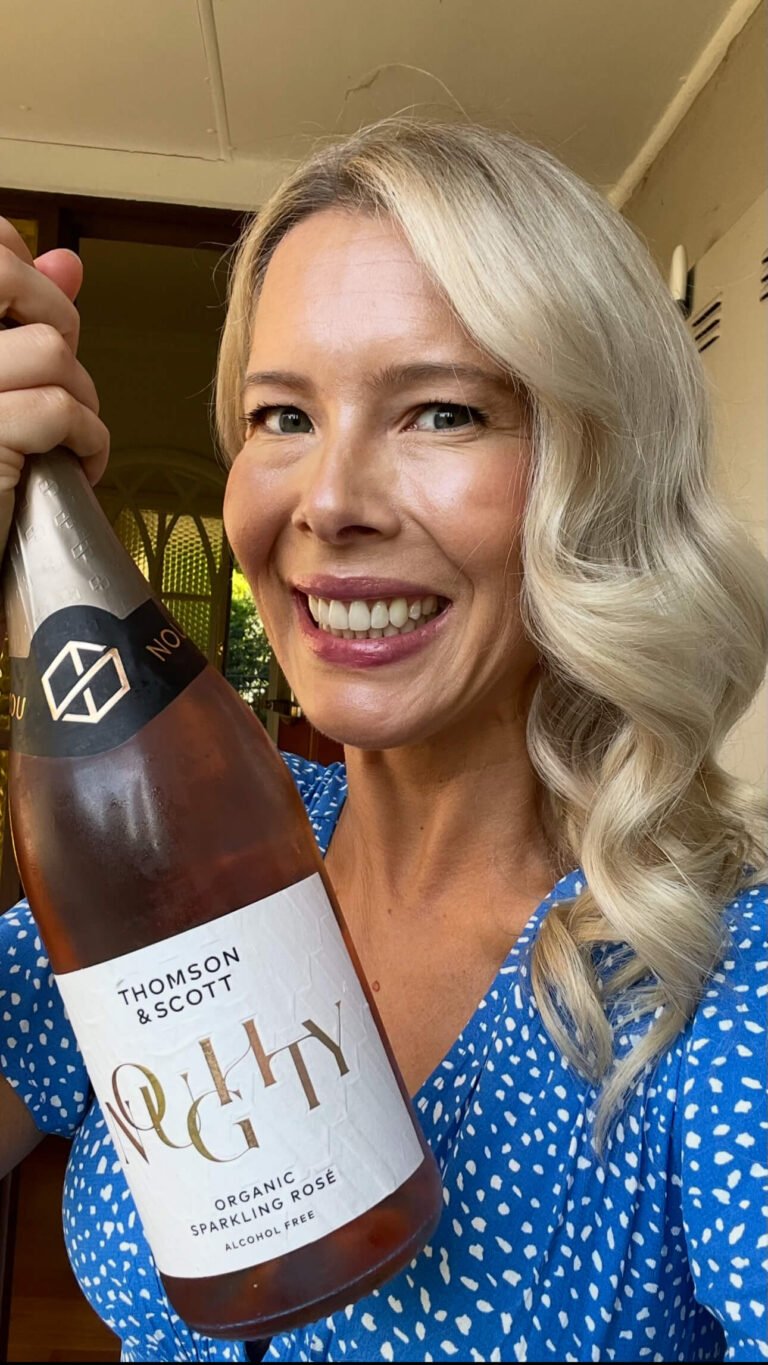 A smiling woman with long blond hair wearing a blue polka dot dress holds up a bottle of Thomson & Scott Noughty organic sparkling rosé. She stands indoors in front of an open door.